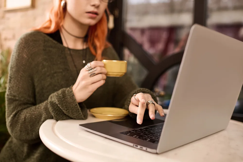 A young woman with long red hair is drinking coffee and looking at her laptop searching for remote jobs that pay weekly